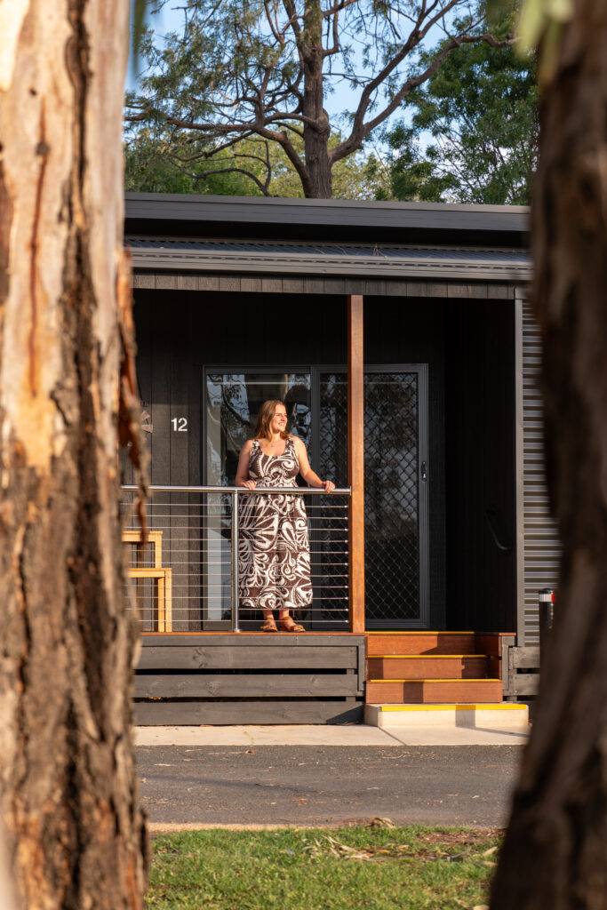Girl standing on a cabin deck between towering gum trees looking into the sunset